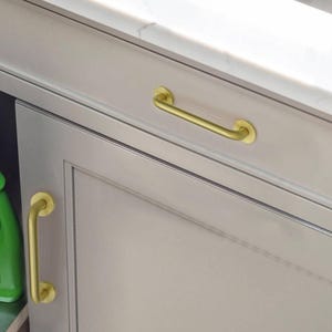 May include: A close-up of a light grey cabinet door featuring a gold-coloured, curved handle. A matching gold-coloured, vertical handle is also visible. The cabinet is set against a white countertop. The handles are made of metal.