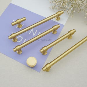 Peut inclure: Ensemble de quincaillerie de meubles dor&eacute;e, comprenant plusieurs poign&eacute;es de barre et un bouton rond. Les poign&eacute;es ont un design cylindrique avec des d&eacute;tails d&eacute;coratifs aux extr&eacute;mit&eacute;s. Le texte "PerfecCrafts" est visible.