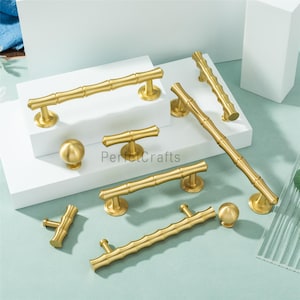 Może przedstawiać: Kolekcja złotych okuć do szafek, w tym uchwytów i gałek. Okucia mają wzór inspirowany bambusem, z teksturowanymi powierzchniami i zaokrąglonymi końcami. Elementy są ułożone na białej powierzchni.