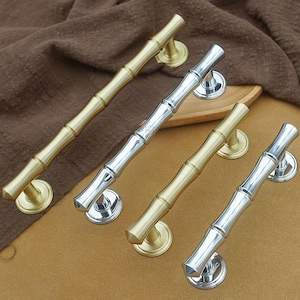 Pode incluir: Quatro puxadores de porta em estilo bambu em tons de ouro e prata. As puxadores estão dispostos diagonalmente, mostrando seu design e ferragens. Os puxadores dourados têm um acabamento quente e latão, enquanto os prateados são polidos.