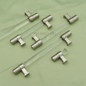 May include: A collection of six cabinet pulls with clear acrylic rods and brushed nickel-colored metal hardware. The pulls are arranged on a light green surface. The text "PerfecCrafts" is visible in the center of the image.