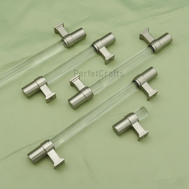 May include: A collection of six cabinet pulls with clear acrylic rods and brushed nickel-colored metal hardware. The pulls are arranged on a light green surface. The text "PerfecCrafts" is visible in the center of the image.