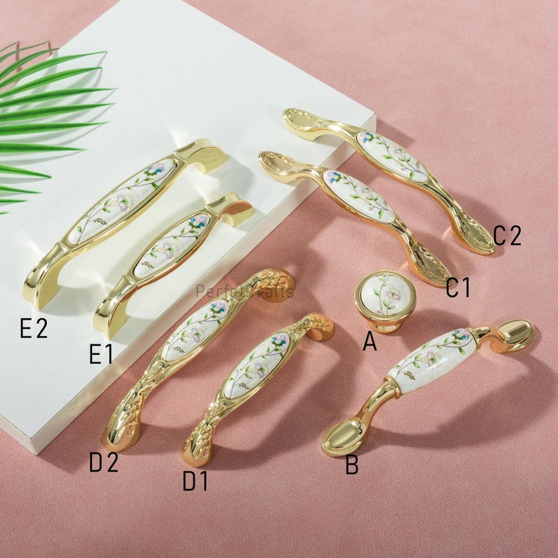 Pu&ograve; includere: Una collezione di ferramenta per mobili color oro, tra cui maniglie e un pomello. La ferramenta presenta un intarsio in ceramica bianca con un design floreale blu, verde e rosa. Varie forme e dimensioni sono esposte su una superficie rosa.