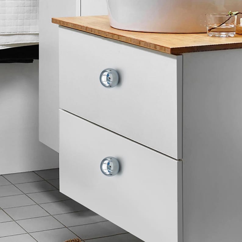 Op de afbeelding: Witte badkamerkast met twee lades, elk met een glanzende, verchroomde knop. De kast heeft een licht houten blad. De vloer is betegeld. De kast staat naast een witte muur.