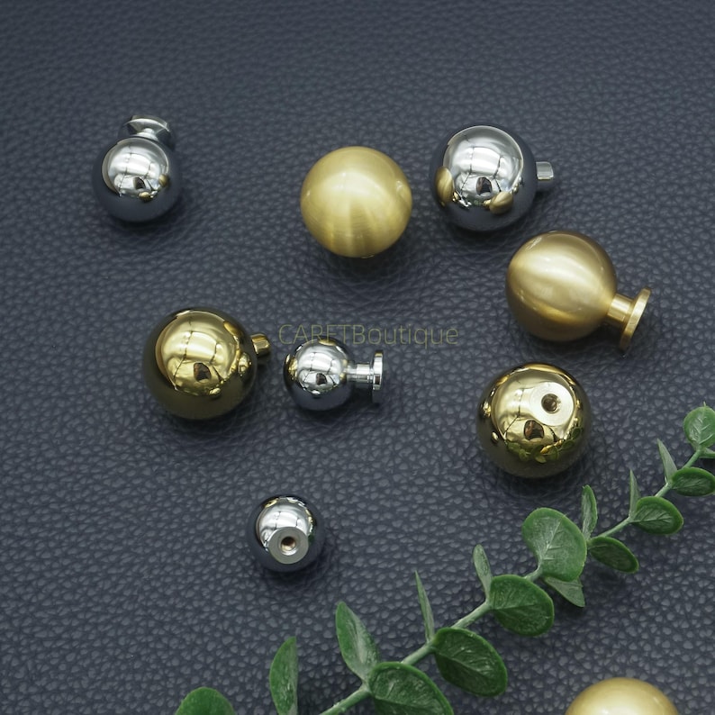 Puede incluir: Un surtido de pomos esf&eacute;ricos para armarios en tonos plateados y dorados pulidos. Los pomos var&iacute;an en tama&ntilde;o y dise&ntilde;o, algunos con forma de bola simple y otros con un v&aacute;stago o base. El hardware se muestra sobre una superficie oscura.