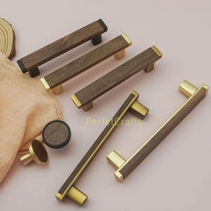 May include: A collection of wooden cabinet pulls and knobs. The pulls are rectangular with a dark wood grain and brass or black accents. One knob is round with a dark wood grain and brass base. The text "Perfectcrafts" is visible.