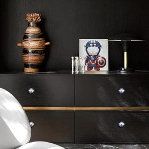Op de afbeelding: Een zwarte dressoir met zilveren knoppen en een gouden accent. Bovenop een keramische vaas met bruine en zwarte strepen, een ingelijste afbeelding van een stripfiguur verkleed als Captain America, een klein zilveren beeld en een zwart-gouden lamp.