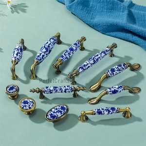 Könnte beinhalten: Eine Auswahl an Keramik-Schrankbeschlägen mit blau-weißem Blumenmuster. Die Beschläge umfassen Knöpfe, Griffe und Züge mit messingfarbenen Metallbasen. Die Artikel sind auf einer hellblauen Oberfläche angeordnet.