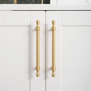 May include: Close-up of two white cabinet doors with vertical, gold-coloured bar pulls. The pulls have a decorative design at the top and bottom. The cabinet doors are clean and modern.