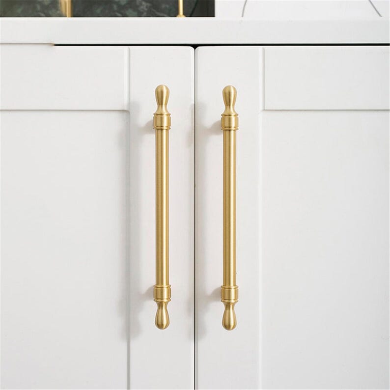 May include: Close-up of two white cabinet doors with vertical, gold-coloured bar pulls. The pulls have a decorative design at the top and bottom. The cabinet doors are clean and modern.