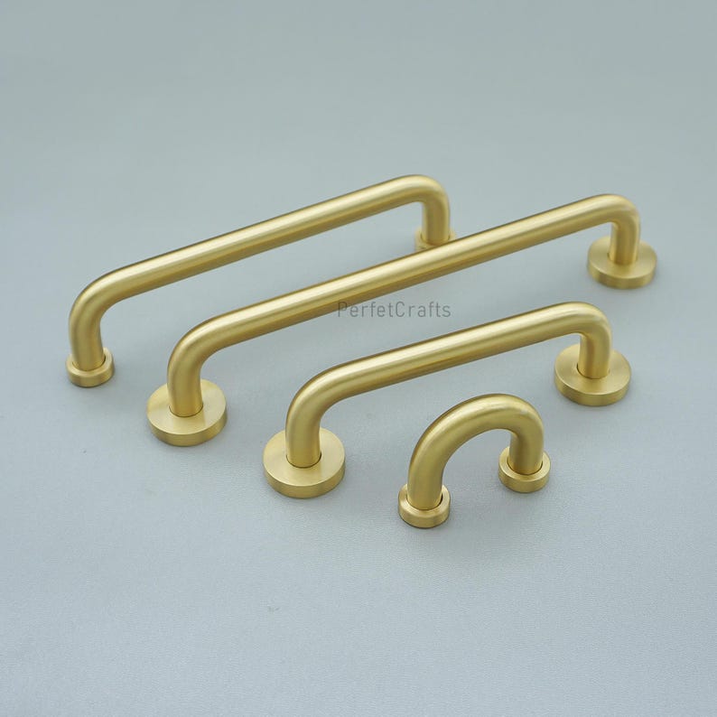 May include: Four brass-coloured cabinet pulls in varying sizes, arranged on a grey surface. Each pull has a curved handle and circular mounting bases. The text "PerfetcCrafts" is visible on the image.