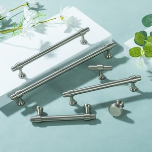 May include: A selection of brushed nickel cabinet handles and knobs. The set includes bar pulls, a T-knob, and a round knob. The hardware is displayed on a white surface with a light blue background, with some greenery and white flowers.
