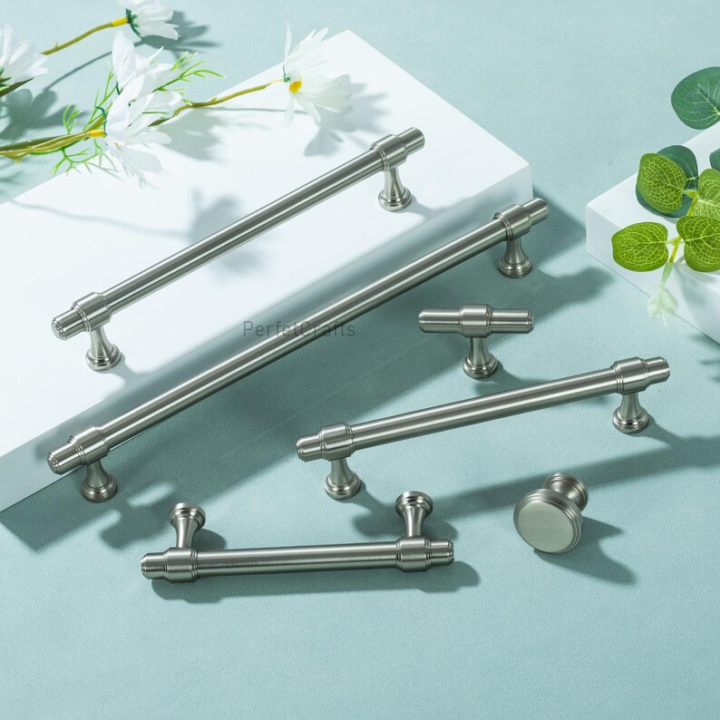 May include: A selection of brushed nickel cabinet handles and knobs. The set includes bar pulls, a T-knob, and a round knob. The hardware is displayed on a white surface with a light blue background, with some greenery and white flowers.