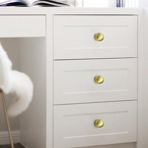 Op de afbeelding: Een witte kast met drie lades, elk voorzien van een ronde, messingkleurige knop. De knoppen hebben een licht getextureerde uitstraling, wat een vleugje elegantie toevoegt aan het minimalistische ontwerp. De kast maakt deel uit van een witte bureau.