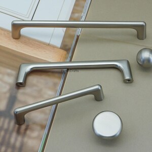 May include: A collection of brushed nickel cabinet hardware, including two bar pulls, a smaller angled pull, and two knobs. The pulls vary in size, with the longest measuring approximately 12 inches. The hardware is displayed on a neutral background.