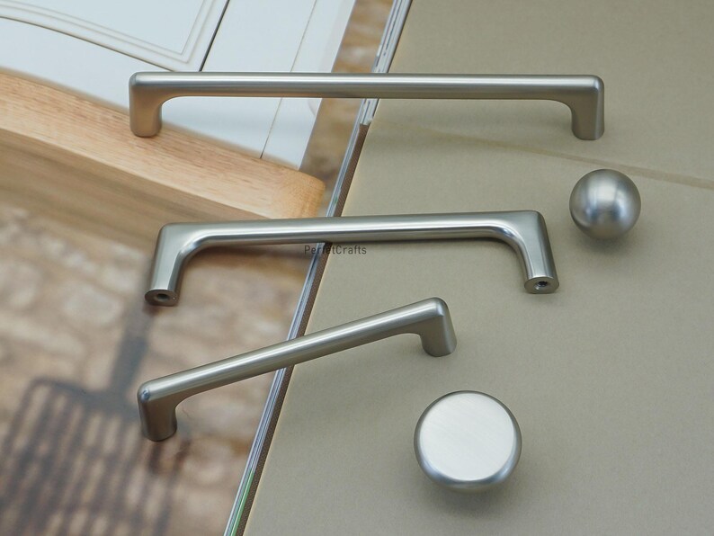 May include: A collection of brushed nickel cabinet hardware, including two bar pulls, a smaller angled pull, and two knobs. The pulls vary in size, with the longest measuring approximately 12 inches. The hardware is displayed on a neutral background.