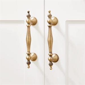 Może przedstawiać: Dwa antyczne mosiężne uchwyty do szafek z ozdobnymi detalami. Każdy uchwyt ma długi, smukły uchwyt z dekoracyjnymi zwieńczeniami i okrągłymi płytkami tylnymi. Okucia są zamontowane na białych drzwiach szafki.