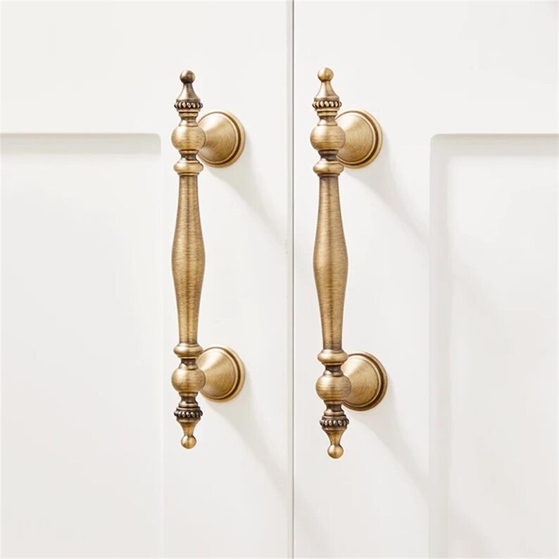 Może przedstawiać: Dwa antyczne mosiężne uchwyty do szafek z ozdobnymi detalami. Każdy uchwyt ma długi, smukły uchwyt z dekoracyjnymi zwieńczeniami i okrągłymi płytkami tylnymi. Okucia są zamontowane na białych drzwiach szafki.