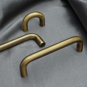 K&ouml;nnte beinhalten: Drei antike Messing-Schrankgriffe in verschiedenen Formen werden auf einer grauen Oberfl&auml;che gezeigt. Die Griffe haben ein gebogenes, r&ouml;hrenf&ouml;rmiges Design mit einem warmen, gealterten Finish. Die Hardware ist wahrscheinlich f&uuml;r M&ouml;bel oder Schr&auml;nke.