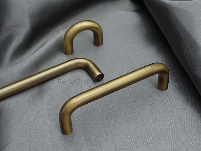 K&ouml;nnte beinhalten: Drei antike Messing-Schrankgriffe in verschiedenen Formen werden auf einer grauen Oberfl&auml;che gezeigt. Die Griffe haben ein gebogenes, r&ouml;hrenf&ouml;rmiges Design mit einem warmen, gealterten Finish. Die Hardware ist wahrscheinlich f&uuml;r M&ouml;bel oder Schr&auml;nke.