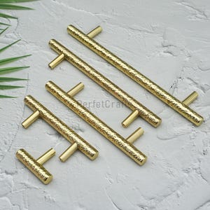 May include: A collection of gold-colored cabinet pulls with a hammered texture. The pulls are arranged diagonally, showcasing their T-bar design. The hardware is likely for furniture or cabinetry.