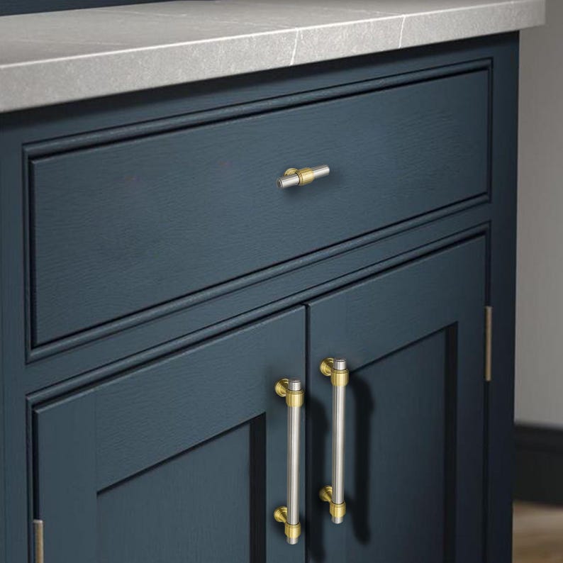 Peut inclure: Gros plan sur un meuble bleu marine avec un plan de travail gris clair. Le meuble a un tiroir avec un bouton dor&eacute; et argent&eacute; et deux portes avec des poign&eacute;es dor&eacute;es et argent&eacute;es assorties. Le meuble est en bois.