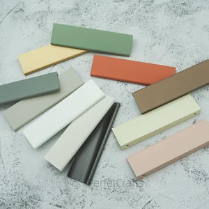 Puede incluir: Una colección de tiradores rectangulares para armarios en varios colores, incluyendo verde, beige, blanco y marrón. Los tiradores están dispuestos sobre una superficie texturizada, mostrando sus diferentes tonos y acabados. El texto "PerfecCrafts" es visible.