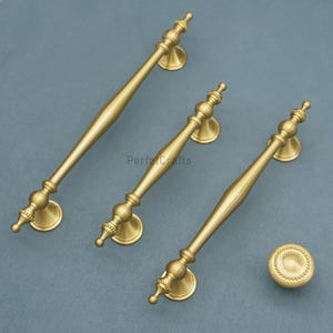 Może przedstawiać: Trzy mosiężne uchwyty do szafek i jeden gałek. Uchwyty mają dekoracyjny design z centralnym uchwytem i ozdobnymi końcami. Gałka jest okrągła z obramowaniem z koralik&oacute;w. Okucia są umieszczone na stonowanym szaro-niebieskim tle.