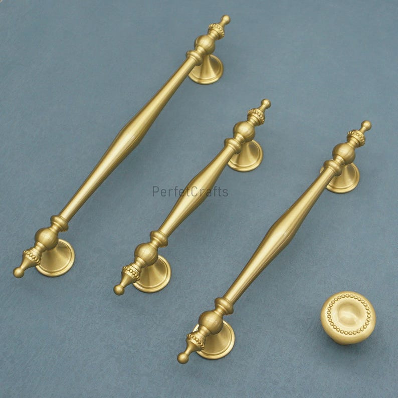 Może przedstawiać: Trzy mosiężne uchwyty do szafek i jeden gałek. Uchwyty mają dekoracyjny design z centralnym uchwytem i ozdobnymi końcami. Gałka jest okrągła z obramowaniem z koralik&oacute;w. Okucia są umieszczone na stonowanym szaro-niebieskim tle.