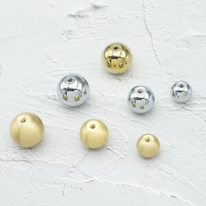 Op de afbeelding: Een assortiment van bolvormige metalen kralen in verschillende maten en afwerkingen. Sommige zijn goudkleurig, andere zilverkleurig met decoratieve uitsparingen. De kralen zijn gerangschikt op een getextureerd wit oppervlak.