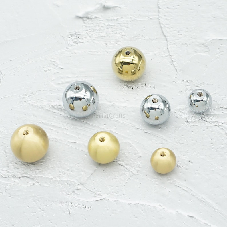 Op de afbeelding: Een assortiment van bolvormige metalen kralen in verschillende maten en afwerkingen. Sommige zijn goudkleurig, andere zilverkleurig met decoratieve uitsparingen. De kralen zijn gerangschikt op een getextureerd wit oppervlak.