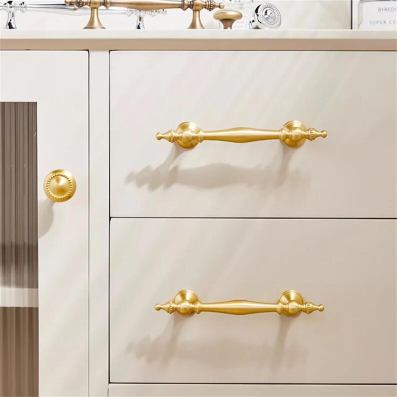 Może przedstawiać: Zbliżenie kremowej szafki ze złotymi okuciami. Szafka posiada dwie szuflady z ozdobnymi, złotymi uchwytami. Pasujący złoty uchwyt znajduje się na drzwiach szafki. Dodatkowe złote okucia są widoczne na g&oacute;rnej p&oacute;łce.