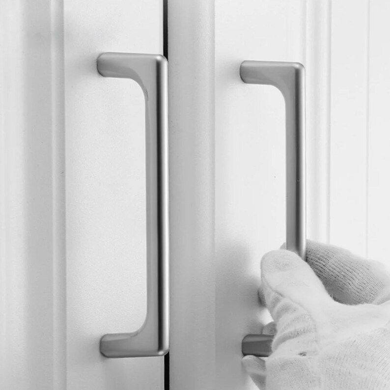 May include: Close-up of two silver cabinet handles. The handles are rectangular with a curved top and bottom. One handle is being held by a gloved hand. The handles are mounted on a white cabinet door.