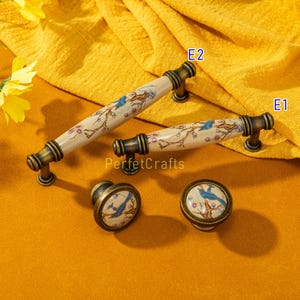 May include: A set of decorative cabinet hardware featuring a bird and floral design. The set includes two handles and two knobs with a cream-colored ceramic finish, accented with bronze-toned metal hardware. The bird is blue, and the flowers are pink.
