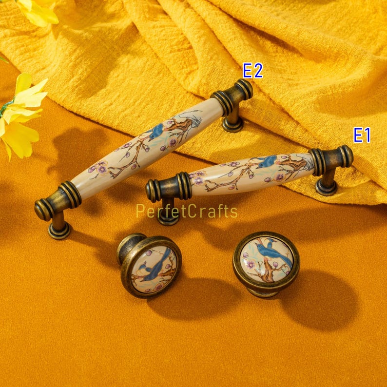 May include: A set of decorative cabinet hardware featuring a bird and floral design. The set includes two handles and two knobs with a cream-colored ceramic finish, accented with bronze-toned metal hardware. The bird is blue, and the flowers are pink.