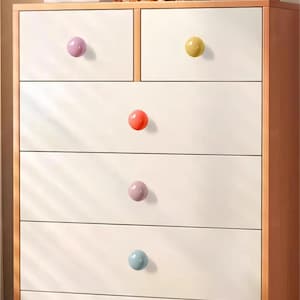 May include: A white chest of drawers with six drawers and colourful round knobs. The knobs are in shades of pink, yellow, orange, and blue. The chest has a light brown wooden frame.