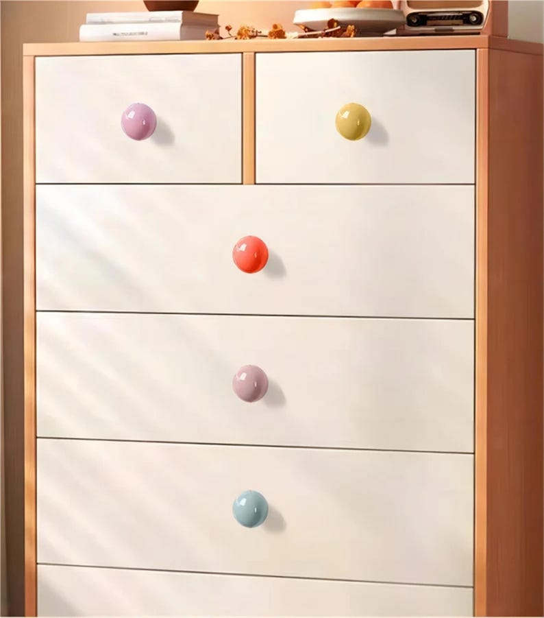 May include: A white chest of drawers with six drawers and colourful round knobs. The knobs are in shades of pink, yellow, orange, and blue. The chest has a light brown wooden frame.