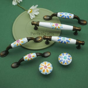 May include: A collection of decorative cabinet hardware, including handles and knobs. The handles have a dark bronze finish with white ceramic centers featuring a floral design in blue, yellow, and pink. The knobs have a similar design.