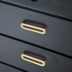 May include: Close-up of a dark blue chest of drawers featuring gold oval drawer pulls. The pulls have a ridged design and are mounted on the drawers. The image highlights the hardware's design and colour against the dark wood.