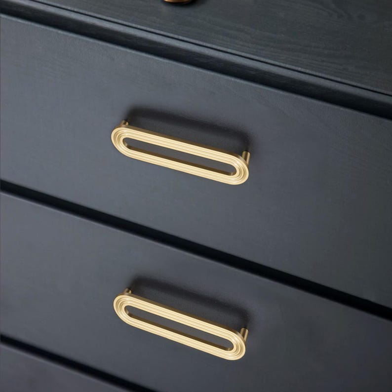 May include: Close-up of a dark blue chest of drawers featuring gold oval drawer pulls. The pulls have a ridged design and are mounted on the drawers. The image highlights the hardware's design and colour against the dark wood.