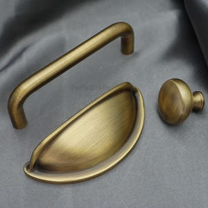 K&ouml;nnte beinhalten: Set aus drei antiken Messing-Schrankbeschl&auml;gen. Enth&auml;lt einen gebogenen Griff, einen Muschelgriff und einen runden Knopf. Die Hardware hat einen warmen, goldenen Farbton und ein geb&uuml;rstetes Finish. Auf grauem Stoffhintergrund.