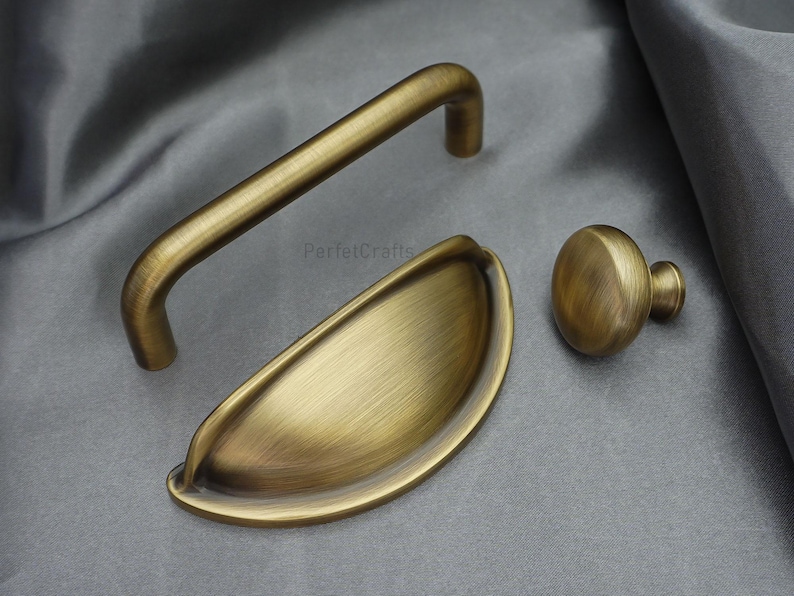 K&ouml;nnte beinhalten: Set aus drei antiken Messing-Schrankbeschl&auml;gen. Enth&auml;lt einen gebogenen Griff, einen Muschelgriff und einen runden Knopf. Die Hardware hat einen warmen, goldenen Farbton und ein geb&uuml;rstetes Finish. Auf grauem Stoffhintergrund.