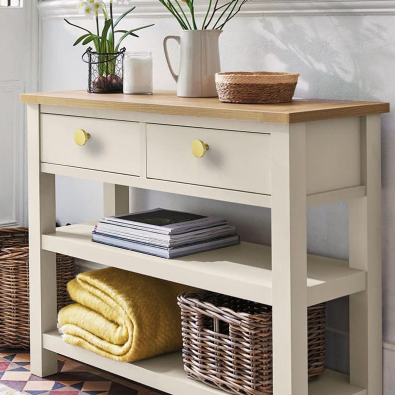 May include: A cream-coloured console table with two drawers and gold knobs. The table has a light wood top and two shelves. A yellow blanket, magazines, and wicker baskets are on the shelves. Decorative items, including flowers, are on top.
