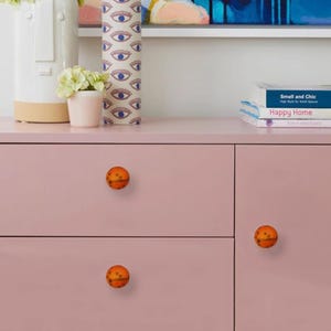 May include: A close-up of a pink cabinet with three orange and brown spherical knobs. The knobs have a textured surface. Decorative items, including a vase and books, are on top of the cabinet.
