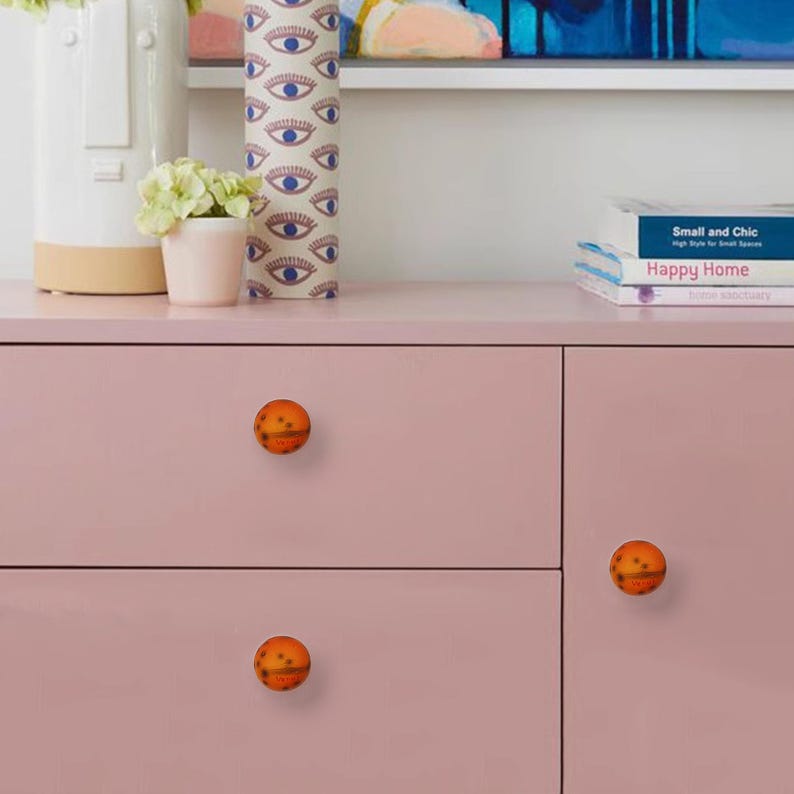 May include: A close-up of a pink cabinet with three orange and brown spherical knobs. The knobs have a textured surface. Decorative items, including a vase and books, are on top of the cabinet.