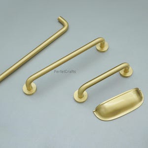 May include: A selection of brass-toned cabinet handles and a cup pull. The image shows various sizes of bar pulls with curved ends and a smooth finish. The hardware is presented on a neutral grey backdrop.