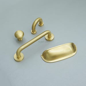 May include: A collection of five brass-toned cabinet fittings. The set includes a knob, two curved handles, a long bar handle, and a rectangular pull. The hardware is shown on a grey surface.