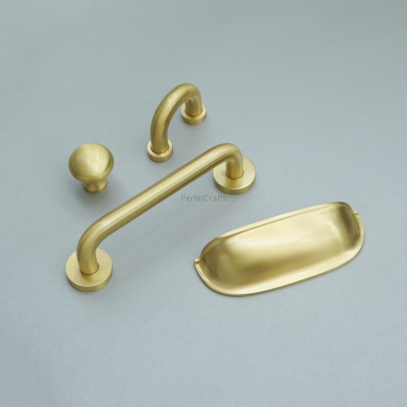 May include: A collection of five brass-toned cabinet fittings. The set includes a knob, two curved handles, a long bar handle, and a rectangular pull. The hardware is shown on a grey surface.