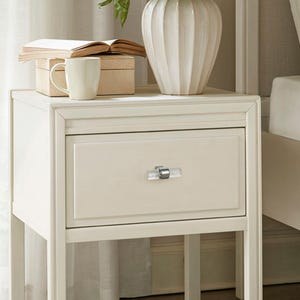 Peut inclure: Table de chevet cr&egrave;me avec un seul tiroir et une poign&eacute;e en forme de cristal. Une tasse blanche, une petite bo&icirc;te en bois et un livre ouvert sont pos&eacute;s sur le dessus, ainsi qu'un vase blanc c&ocirc;tel&eacute;. Le chevet fait partie d'un ensemble de meubles de chambre.