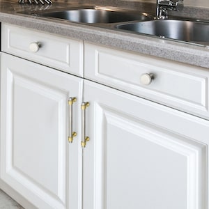 Peut inclure: Des armoires de cuisine blanches avec des ferrures argent&eacute;es et dor&eacute;es. Les armoires pr&eacute;sentent un design &agrave; panneaux sur&eacute;lev&eacute;s et sont accentu&eacute;es de boutons argent&eacute;s et de barres dor&eacute;es et argent&eacute;es. Le comptoir est gris mouchet&eacute;.